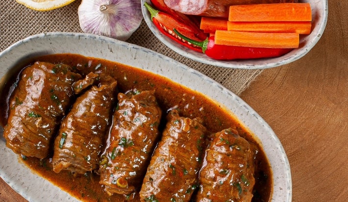 Em cima de uma mesa, refratário com bifes suculentos enrolados com molho de tomate, decorado com ervas.