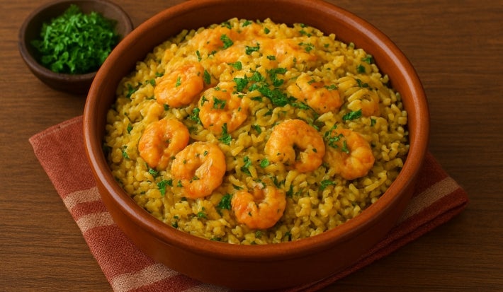 Fotografia de um recipiente marrom de vidro fundo, médio, sobre um pano dobrado de tom rosa e branco apoiado em uma mesa de madeira. Dentro do recipiente tem arroz paraense com camarões por cima e um pouco de cheiro verde salpicado.