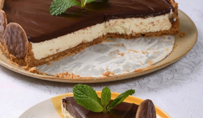 Em cima de uma mesa, prato redondo com fatia de torta de biscoitos calipso, com creme branco e ganache de chocolate.