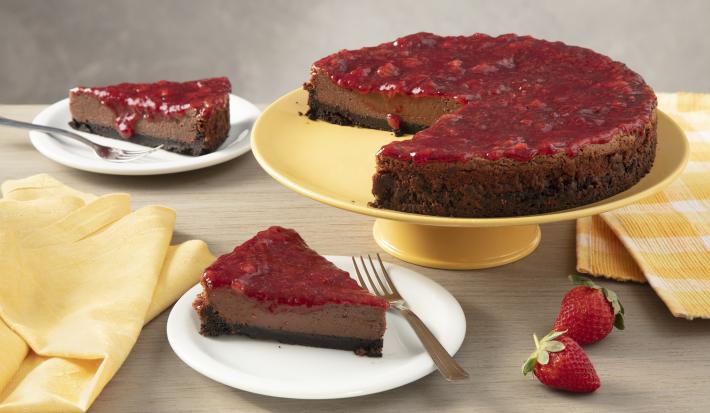 Fotografia em tons de amarelo em uma bancada de madeira com um suporte para bolo amarelo ao centro e a torta cheesecake de chocolate em cima. Ao lado, um paninho xadrez amarelo e dois pratos brancos com duas fatias da cheesecake de chocolate em cada um.
