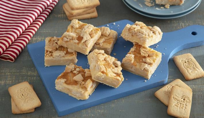 Foto em tons de bege da receita de doce de micro-ondas de biscoito passatempo servida em diversas porções sobre uma tábua azul com algumsa unidades de biscoito passatempo leite em volta. Ao fundo um paninho listrado vermelho e branco e um prato azul