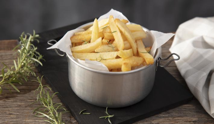 Imagem de um pote de alumínio forrado com papel e batatas fritas dentro. Ao lado, ramos de alecrim.