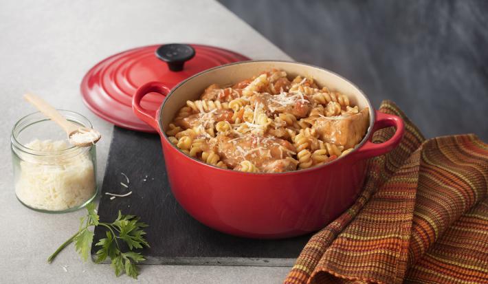 Fotografia de uma panela vermelha aberta com macarrão parafuso, frango e queijo parmesão ralado em cima e ao lado está a tampa da panela vermelha com ponta preta, um guardanapo listrado em tons terrosos e uma pode de vidro transparente com queijo ralado.