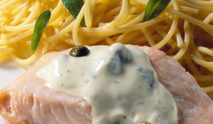 Uma mesa azul com bolinhas brancas vista de cima, um prato quadrado branco com macarrão espaguete, um pedaço de salmão com molho branco por cima.