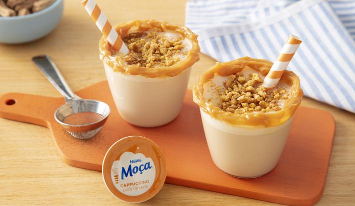 Fotografia em tons de laranja em uma bancada de madeira de cor marrom. AO centro, uma mini tábua de madeira contendo as duas bebidas. Ao lado, 1 cápsula de Nescafé e ao fundo, um pano azul listrado.
