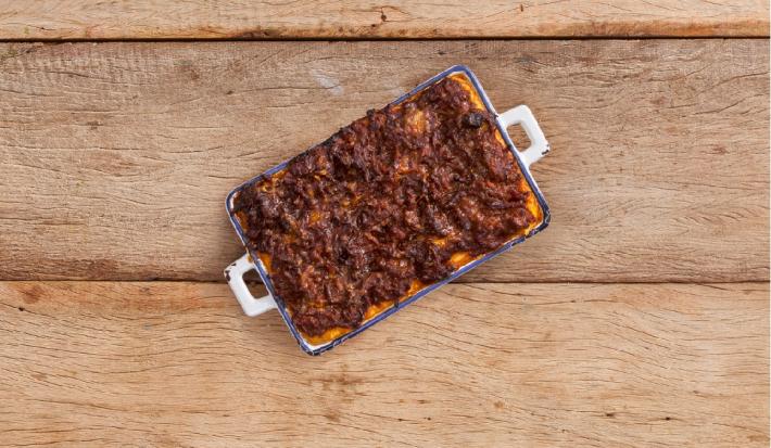 Fotografia em tons de marrom em uma bancada de madeira rústica com um recipiente azul com alças brancas laterais e a polenta cremosa coberta com molho de carne dentro do recipiente.