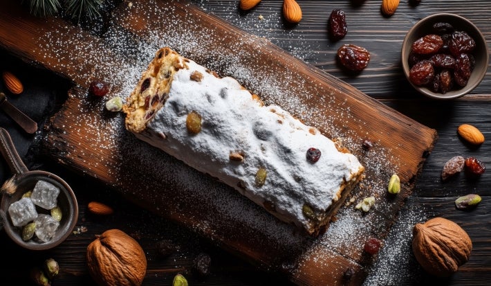 Fotografia de um bolo em uma tábua rústica, coberto com açúcar de confeiteiro. Acompanhado de nozes, amêndoas e um bowl de passas, a composição destaca tons quentes e texturas naturais