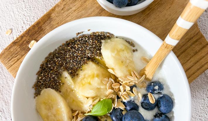 Foto da receita de bowl plant-based. Observa-se um bowl branco com mirtilos, banana, aveia e chia sobre uma tábua de madeira.