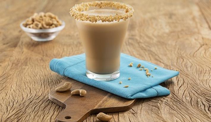 Foto da Receita de Cappuccino Gelado de Caju. Observa-se um copo transparente de vidro decorado com castanha de caju no topo sobre um guardanapo azul e uma pequena tábua de madeira. Atrás, tem um potinho com castanhas de caju.
