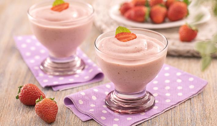 Foto em tons de vermelho da receita de sorvetinho cremoso servida em duas taças de vidro redondas decoradas com um paninho rosa com bolinhas brancas embaixo. Ao lado e ao fundo há algumas unidades de morango além de outras espalhadas em um prato ao fundo