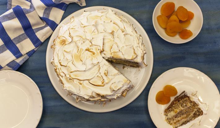 Fotografia em tons de azul e laranja em uma bancada azul, com um pano xadrez azul, um prato branco redondo com a torta de nozes em cima dele. Ao lado, um prato menor com a fatia da torta e damascos ao lado dele.
