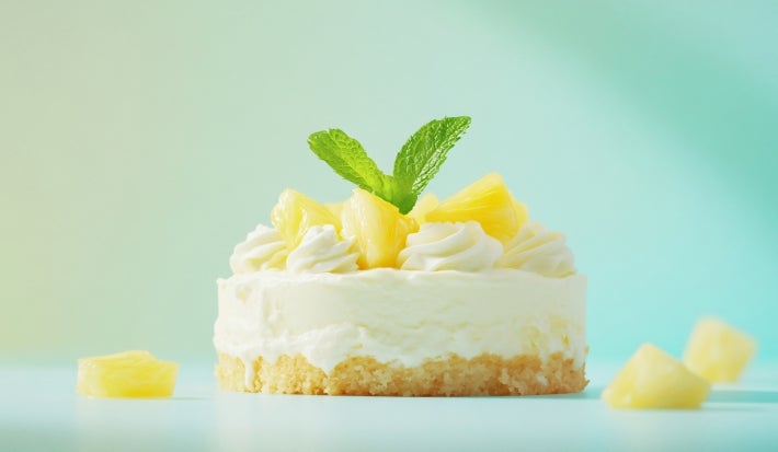 Fotografia em tons claros de uma mini torta de abacaxi decorada com duas folhas de hortelã, o fundo tem tom verde água e tem alguns cubinhos de abacaxi em volta da torta.