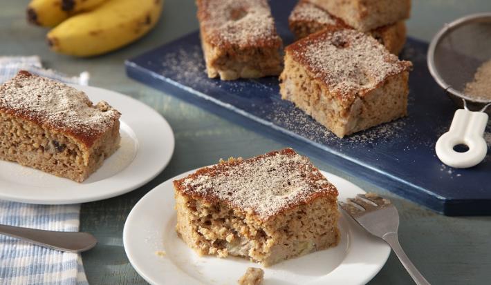 Foto da receita de bolo de banana com farinha láctea aveia servida em duas porções dispostas sobre pratos de porcelana brancos com uma régua azul atrás com mais pedaços do bolo em cima. Ao fundo ainda há duas bananas e uma peneira com canela em pó