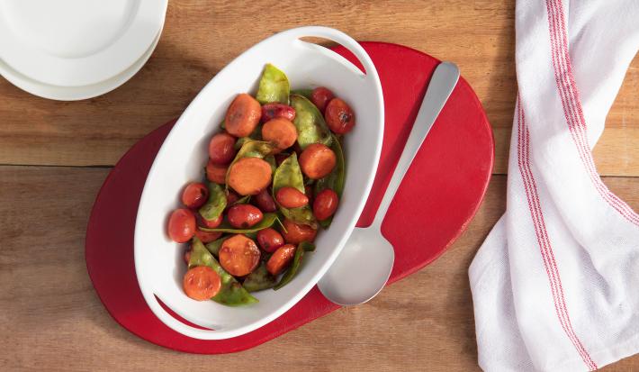 Fotografia em tons de vermelho em uma bancada de madeira, um pano branco com listras vermelhas, um suporte de panela vermelho, um recipiente branco oval com os legumes glaceados dentro.