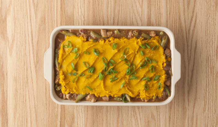 fotografia em tons de marrom e amarelo de uma bancada marrom vista de cima, contém um recipiente branco com alças com o pedaços de carne, purê de abóbora e cebolinhas por cima