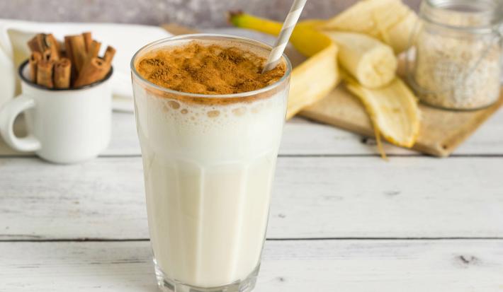 Fotografia de um copo de vidro com uma vitamina de banana com canela polvilhada e um canudo branco e cinza dentro do copo. Está sobre uma mesa de madeira branca, ao fundo tem paus de canela em um copinho branco e metade de uma banana aberta.