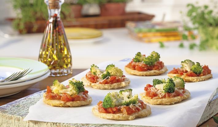 Fotografia em tons de amarelo e verde em uma bancada de madeira, com uma mesa arrumada para um jantar. Ao centro, uma tábua de madeira com um papel manteiga e várias mini pizzas com tomate e brócolis em cima dele.