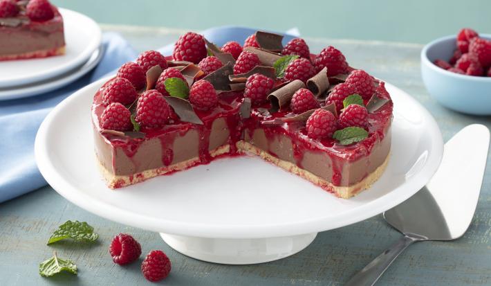 Fotografia em tons de vermelho em uma bancada de madeira azul, um pano azul claro, um suporte para bolo branco com a cheesecake de chocolate com calda de framboesa em cima do suporte, decorado com raspas de chocolate e framboesas.