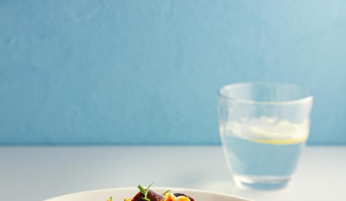 Fotografia de macarrão tipo parafuso com molho de tomate e berinjela dentro de um prato branco de vidro fundo. Ao redor da pasta tem um garfo e um copo de vidro com água e limão. O prato está sobre uma mesa branca.