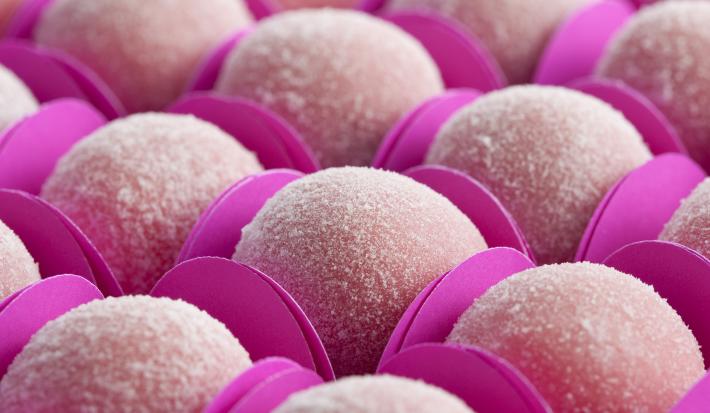 Fotografia em tons de rosa, com docinhos rosa claro na forma de bolinhas, envoltos em açúcar e dentro de forminhas de papel cartão na cor pink.