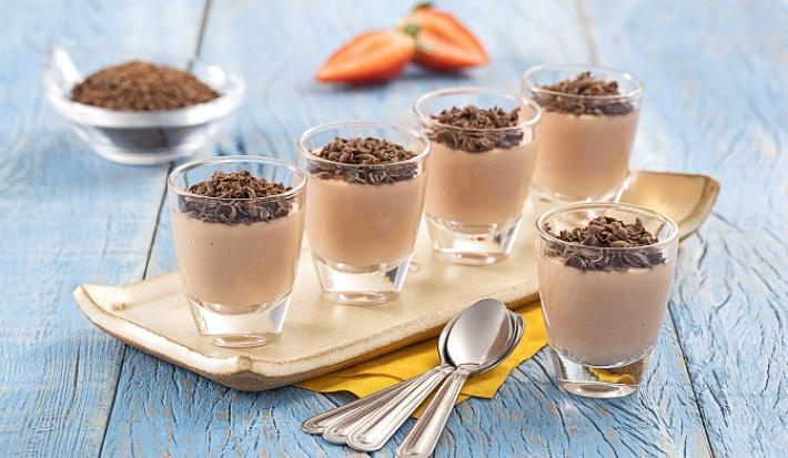 Foto de Receita de Brigadeiro de Morango com Nibs de Cacau. Observa-se uma peça de cerâmica retangular com cinco copinhos de vidro transparentes sob uma madeira azul clara.