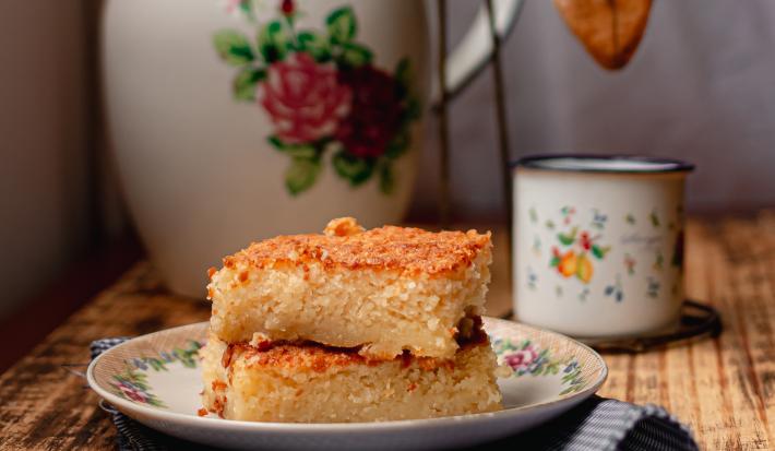 Foto da receita de Bolo de Arroz Cozido. Observa-se 3 fatias retangulares de bolo empilhadas e, atrás, um cafézinho para acompanhar.