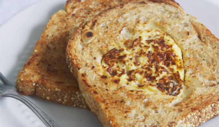 Fotografia em tons de bege em uma mesa de madeira clara com um prato branco raso e duas fatias de pão integral com a omelete dentro dele.