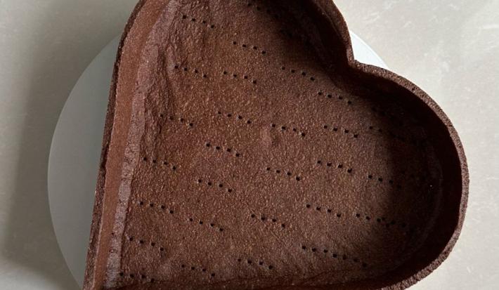 Fotografia em tons claros de uma massa de torta sabor chocolate em um prato branco raso sobre uma mesa branca.