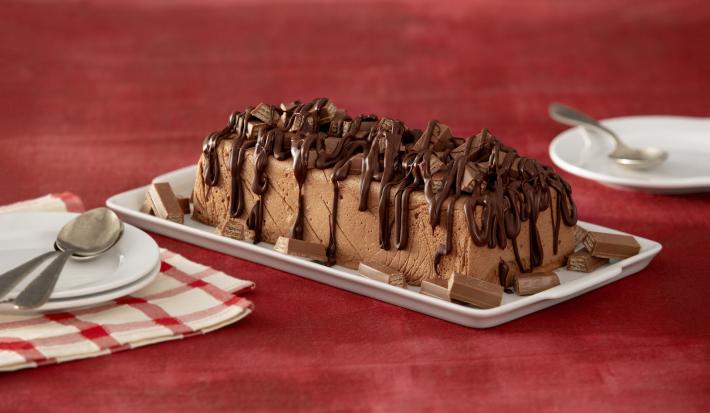 Fotografia em tons de chocolate e vinho, ao centro régua branca contendo torta de chocolate coberta e decorada com chocolate, ao lados pratos com colheres, dois deles sobre guardanapo branco com listras vermelhas, tudo sobre bancada na cor vinho.