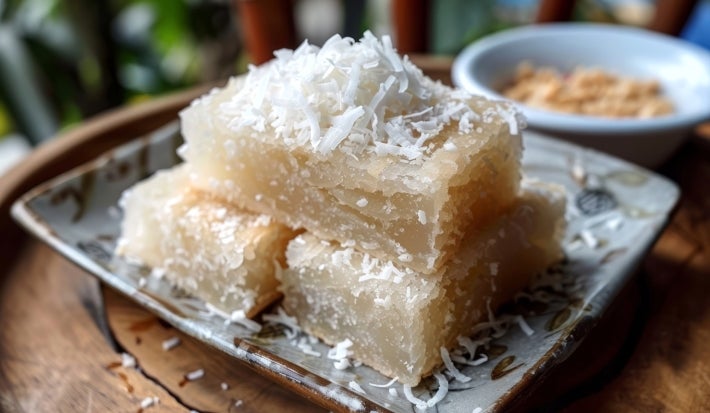 Fotografia de um prato de sobremesa tradicional, com pedaços retangulares de bolo de tapioca cobertos com coco ralado em um prato decorativo. O fundo desfocado sugere um ambiente natural.
