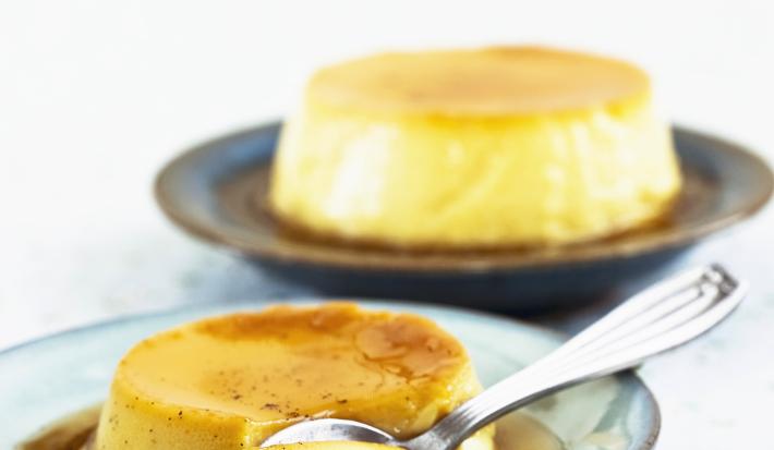 Fotografia de um flan de iogurte e maracujá com calda de maracujá com uma colher de sobremesa cortando um pedaço sobre um prato de tom verde. Ao fundo, outro flan, sobre uma mesa branca.