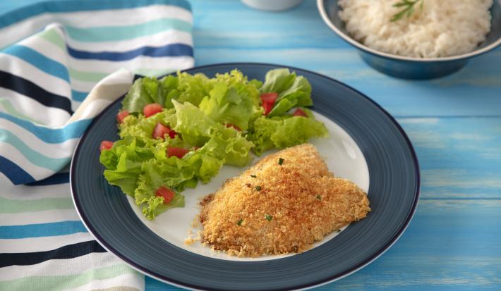 Foto da receita de Filé de Tilápia Copacol com Crosta de Parmesão, vista de cima, em um prato redondo servido com salada, em uma bancada azul com talheresl