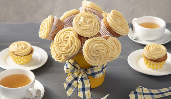 Fotografia em tons de amarelo em uma bancada cinza com um vaso de buquê de cupcake ao centro com um laço xadrez em amarelo e azul. Ao lado, xícaras brancas com chá e pratos brancos com um cupcake em cada um.