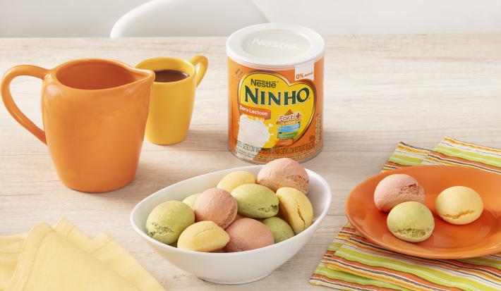 Fotografia em tons de branco e laranja de uma bancada branca com um recipiente branco oval, sobre ele pães de queijo. Ao lado um paninho listrado com um prato laranja com pães de queijo, uma lata de NINHO zero lactose, uma xícara e uma jarra laranjas.