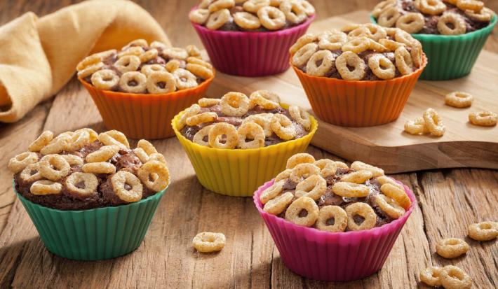 Fotografia em tons de laranja, verde, amarelo e rosa em uma bancada de madeira clara, um paninho amarelo, uma tábua de madeira, e várias forminhas de doces coloridas com o cupcake de chocolate cobertos com cheerios.