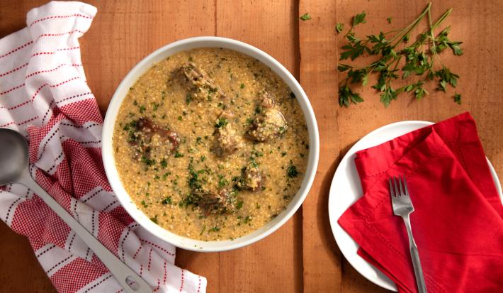 de uma bancada de madeira vista de cima contém um recipiente branco e redondo ao centro com pedaços de carne e canjiquinha. De um lado um prato redondo e branco e por cima uma pano vermelho com um garfo para servir, e do outro um pano branco e rosa