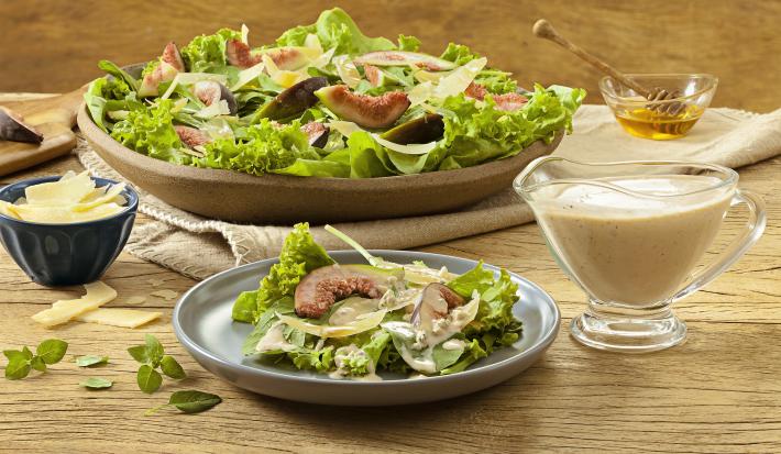 Fotografia em tons de verde em uma bancada de madeira com um prato de madeira grande com salada de alface, figo e queijos. Mais à frente, um prato menor com uma porção pequena da salada. Ao lado, um potinho com molho, mel e queijo parmesão.