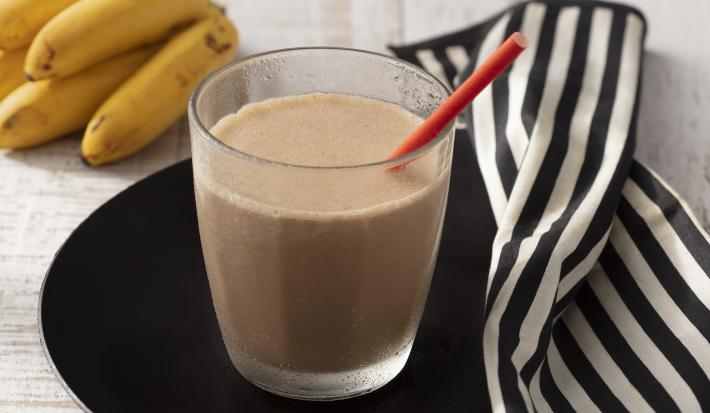 Fotografia em tons de branco e preto de uma bancada de madeira branca com um prato preto, sobre ele um copo de vidro com a bebida cremosa e um canudo vermelho. Ao lado um paninho listrado branco e preto e um cacho de banana.
