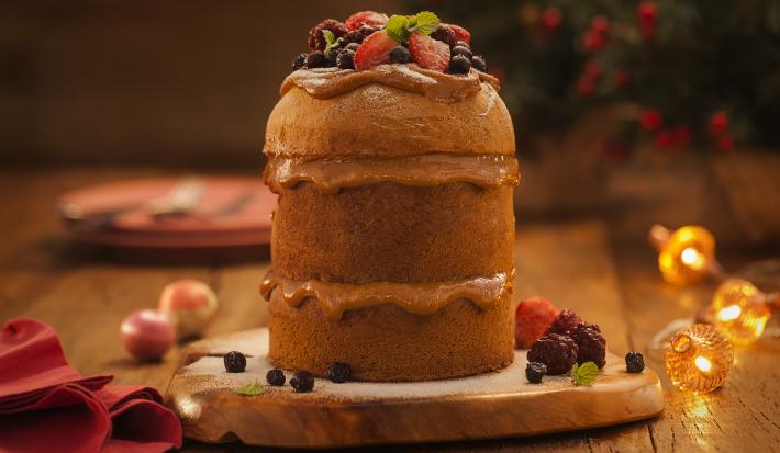 Foto de um panettone cortado em três camadas, com um recheio em tom de marrom claro. Ele está sobre uma tábua de madeira há decorações de natal