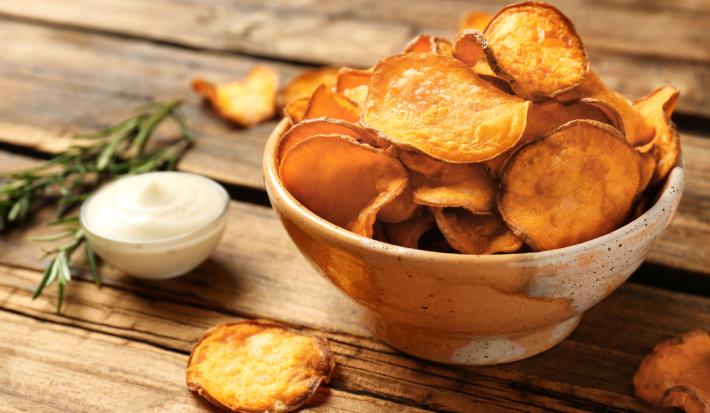Fotografia em tons de marrom com um prato fundo de cerâmica ao centro. Em cima do prato existe uma porção de chips de batata doce feitos na Air Fryer