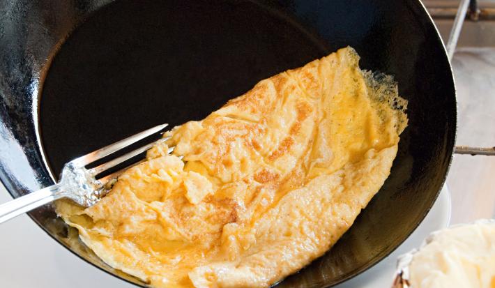Fotografia de uma omelete dobrada em uma frigideira funda preta que está inclinada para colocar o ovo no prato, e um garfo segundo a omelete. Ao fundo, ovos sobre uma mesa branca. Ao lado do prato que vai receber a omelete, um pano de mesa vermelho.