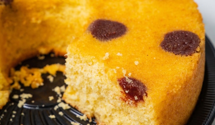 Fotografia de um close de um bolo amarelo com uma fatia removida, mostrando sua textura macia e úmida. O bolo está em uma bandeja circular preta, com pedaços de goiabadas, destacando sua aparência apetitosa.