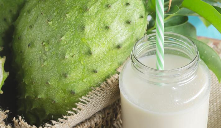 Fotografia em tons de verde e branco de uma bancada vista de cima, contém um copo transparente com suco de graviola e dentro um canudo verde e branco e ao lado uma graviola dentro de um cesto marrom quadrado