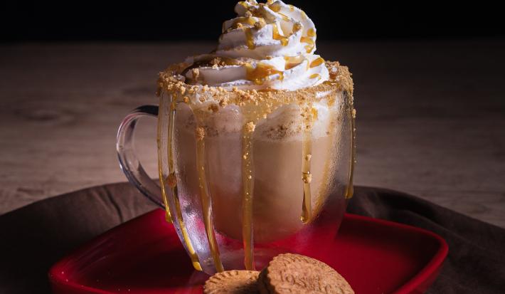 Foto da receita de cappuccino caramelo salgado servida em dois copos de vidro sobre uma mesa de madeira com uma mão polvilhando caramelo salgado
