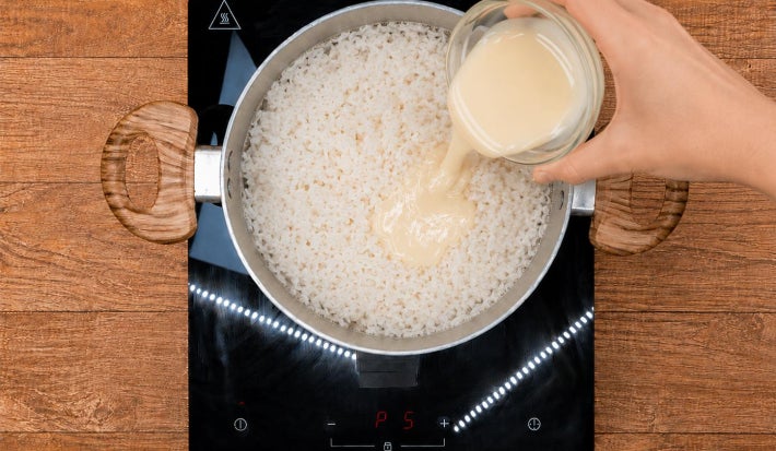 Mão despejando Leite Moça de tigela de vidro em panela com arroz cozido sobre fogão portátil preto