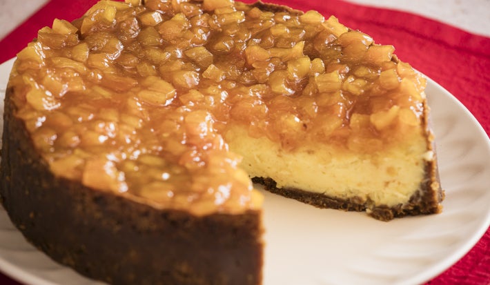Foto da Receita de Cheesecake de Queijo Brie com Calda de Damasco e Laranja, servida em um prato branco, com uma fatia faltando, sobre um pano vermelho