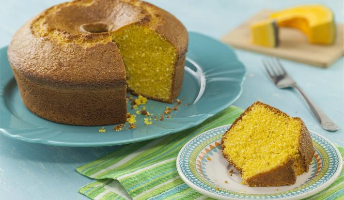 Fotografia em tons de azul de uma bancada azul com um prato azul, sobre ele um bolo de abóbora. Ao lado um paninho verde listrado com um prato redondo pequeno com uma fatia do bolo. Ao fundo uma tábua com uma fatia de abóbora.