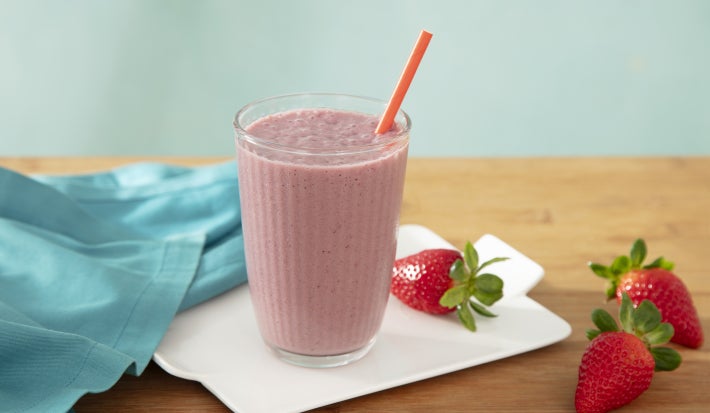 Foto da receita de shake de açaí com novasource proline servida em um copo de vidro alto sobre uma louça branca em cima de uma mesa de madeira com morangos