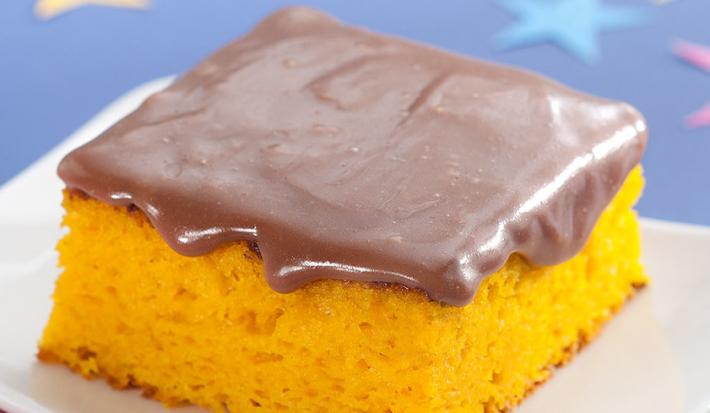 Fotografia em tons de azul, laranja e marrom de uma bancada azul, sobre ela um prato branco quadrado com uma fatia de bolo de cenoura com cobertura de chocolate. Ao lado um garfo verde. Ao fundo uma estrela azul e uma rosa.