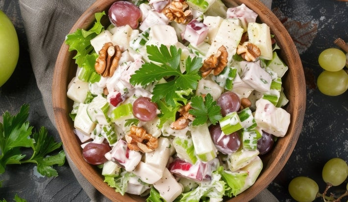Fotografia de uma salada vibrante em uma tigela de madeira. A mistura inclui cubos de batatas cozidas, maçã-verde, uvas roxas e nozes, coberta com um molho cremoso e decorada com salsa, destacando frescor e texturas harmoniosas.